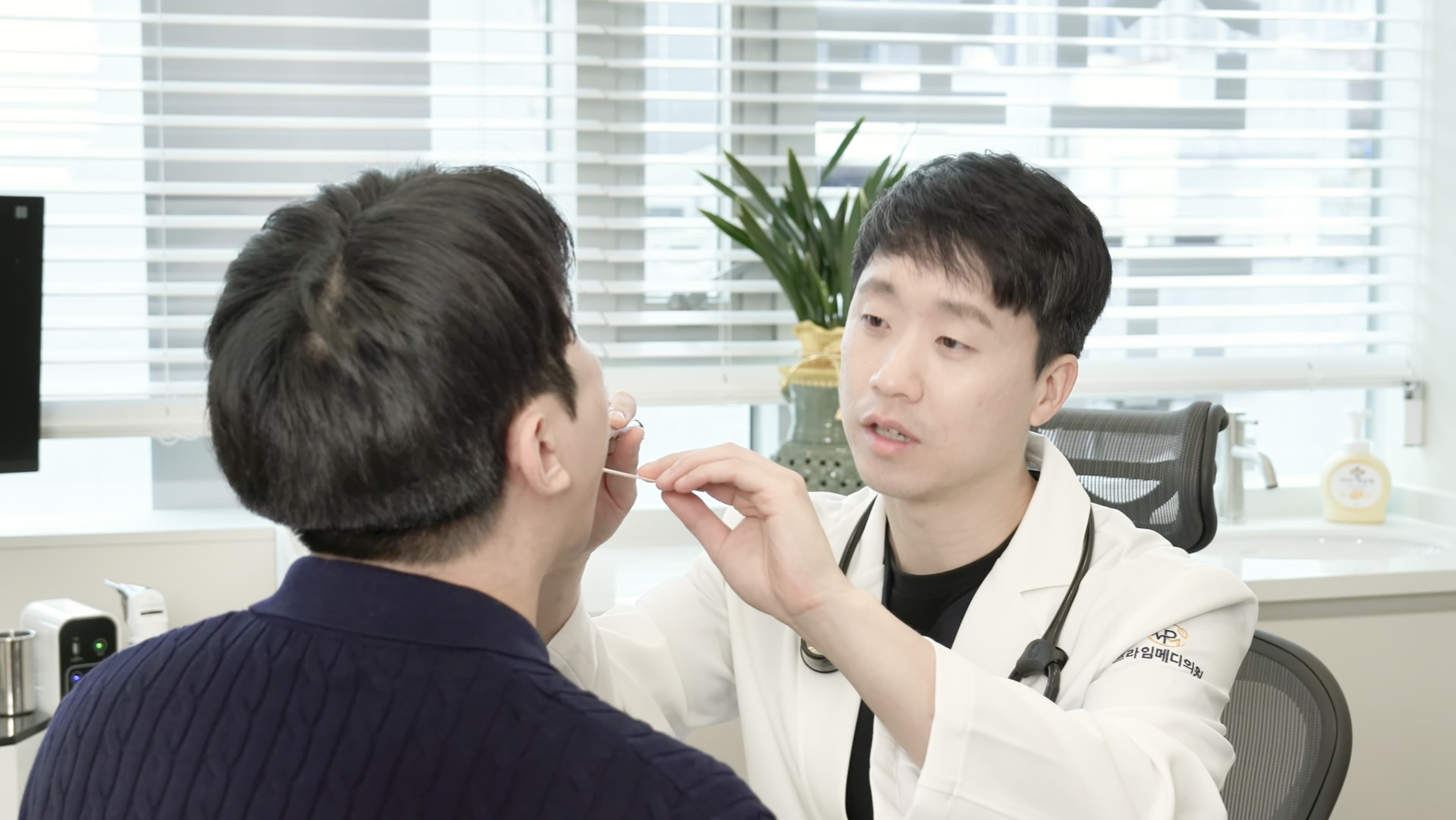 안양프라임메디의원 가정의학과 전문의 문다운 원장이 환자의 얼굴과 귀 주변을 가까이에서 살펴보며 세심하게 진찰하는 모습으로, 밝은 채광이 드는 진료실 분위기를 함께 보여준다.
