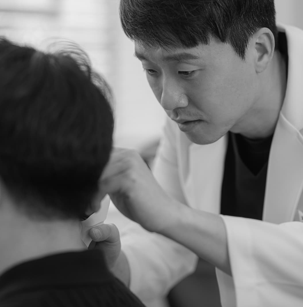 가정의학과 전문의 문다운 원장이 환자의 목과 어깨 부위를 두 손으로 짚어가며 세심하게 촉진하는 모습을 담은 흑백 사진. 신중하고 전문적인 진료 철학을 시각적으로 전달한다.